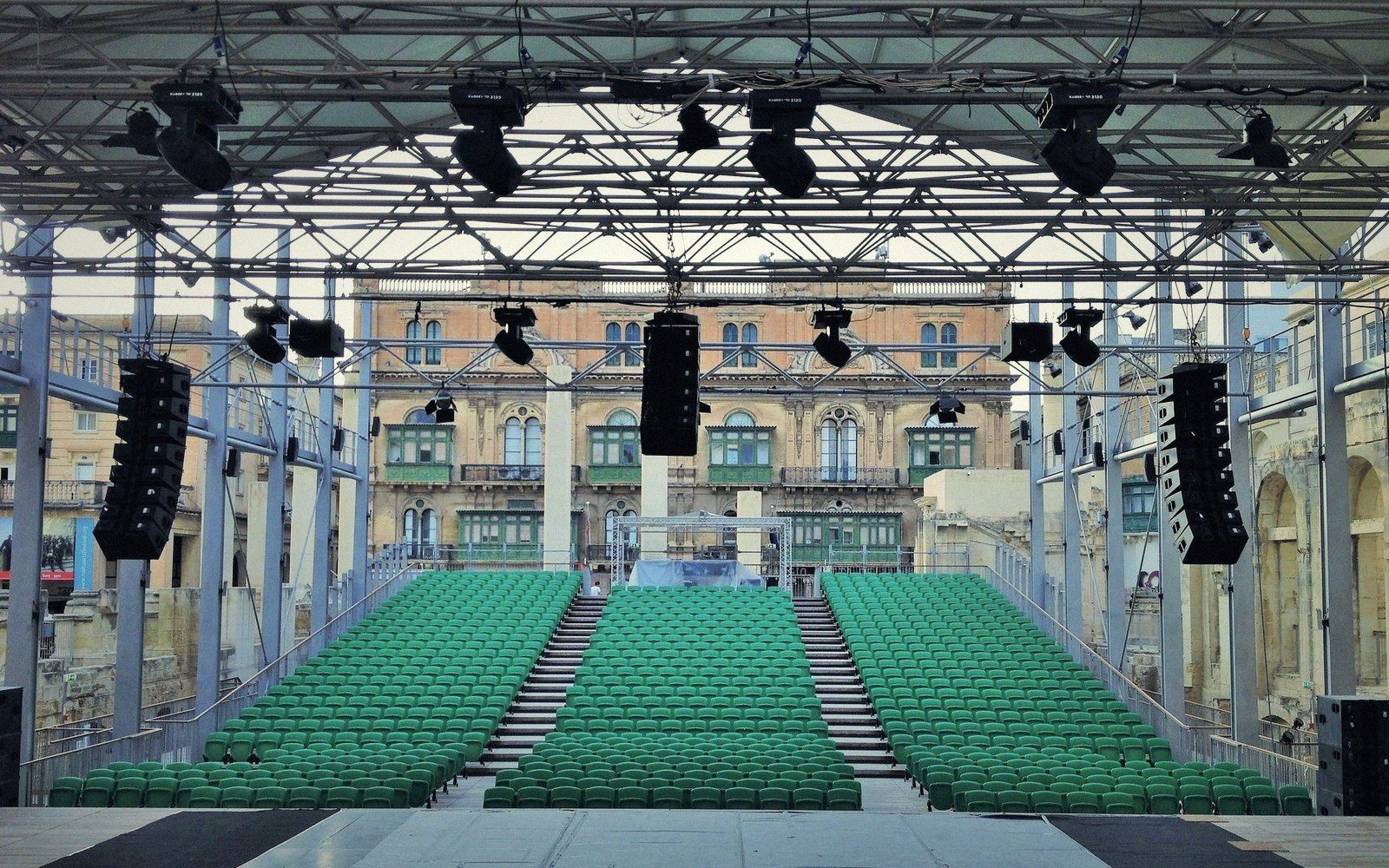 Rows of green seats face a stage under a rigged canopy of lights facing a historic facade