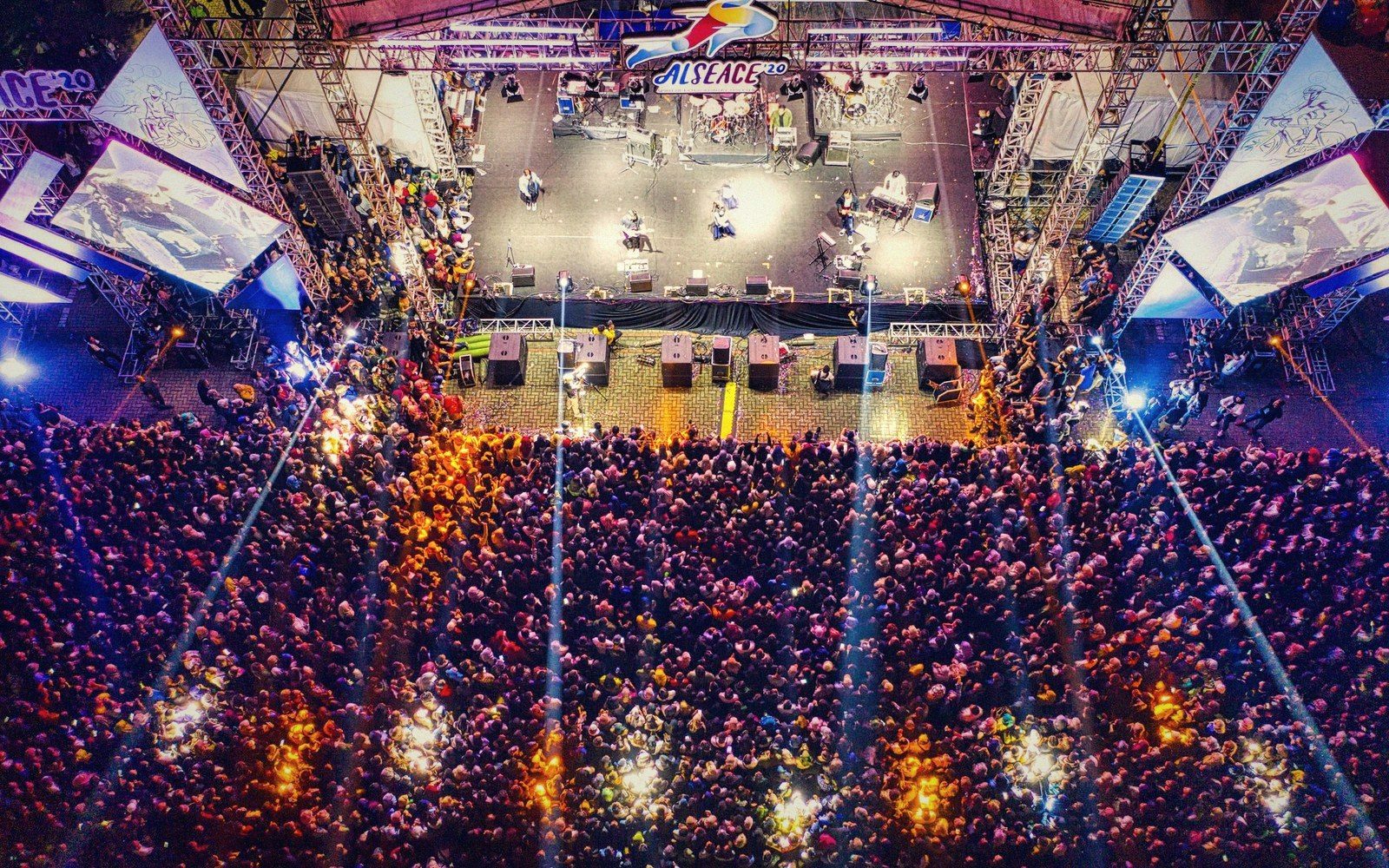 Aerial view of a crowded outdoor concert with a lit stage and screens on either side